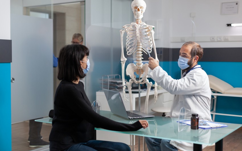 orthopedic-specialist-explaining-bones-injury-on-human-skeleton-to-woman-in-medical-cabinet-physician-pointing-at-spinal-cord-on-osteopathy-model-to-show-anatomy-structure-and-diagnosis.jpg
